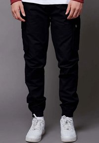 Pantaloni cargo neri con polsini elasticizzati, tasche laterali e dettaglio del logo. Indossati con sneakers bianche, caratterizzate da una trama liscia e resistente.