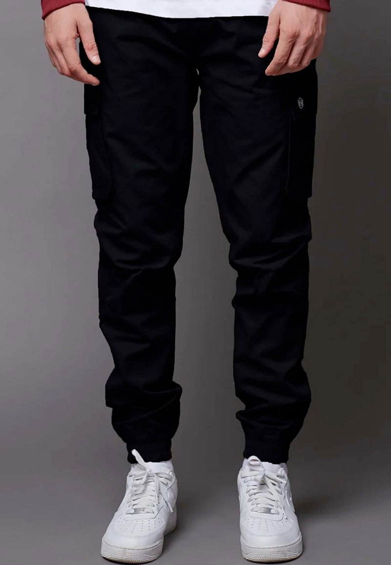 Pantaloni cargo neri con polsini elasticizzati, tasche laterali e dettaglio del logo. Indossati con sneakers bianche, caratterizzate da una trama liscia e resistente.