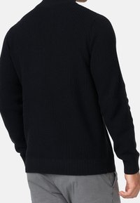 Homme portant un pull en maille texturée noir et un pantalon gris clair, vu de dos sur un fond uni.