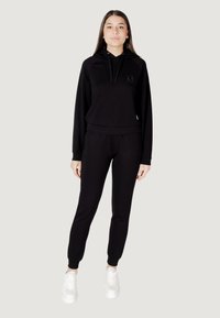 Czarna bluza z kapturem z małym logo, w zestawie z czarnymi dresami, obie z miękkiego, teksturowanego materiału. Białe trampki.