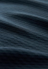 Textuur donkerblauwe stof met een subtiel geometrisch patroon, met zachte plooien en een gladde oppervlakte. Het materiaal lijkt lichtgewicht en ademend te zijn.