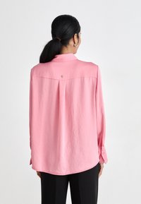 Rosa langermet skjorte med krage, med en bakpleat og en liten logodetalj. Teksturert stoff og knappede mansjetter.