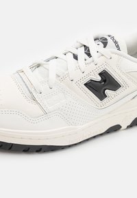 Vita lädersneaker med perforerade detaljer, svart logotyp, texturerad sula och platta snören. Style-nummer "550" präglat på sidan.