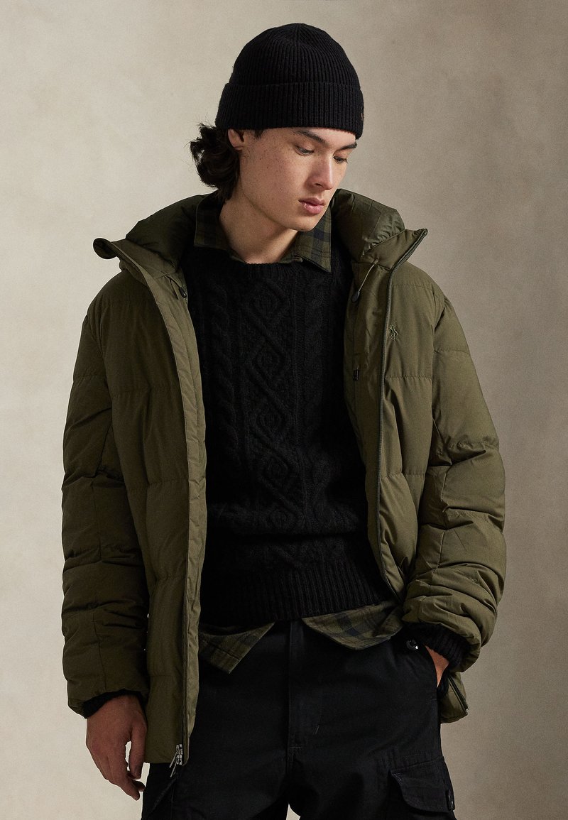 Casaco almofadado verde-oliva com uma camisola preta texturada por baixo. A modelo veste um gorro preto em malha canelada e uma camisa verde aos quadrados.