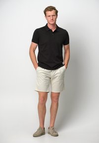 Svart pikétröja med korta ärmar, beige shorts och beige sneakers. Modell står med händerna i fickorna mot en enkel bakgrund.
