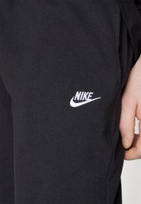 Černé tepláky vyrobené z bavlněné směsové látky s hladkou texturou, se bílým vyšitým logem Nike na boku.