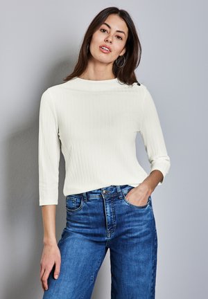 Crèmekleurige ribbeltrui met lange mouwen, een bootneck en driekwartmouwen, gecombineerd met blauwe denim jeans. Opvallende knoopdetails op de schouder.