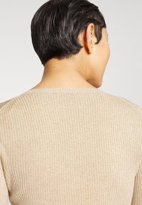 Personne aux cheveux courts noirs portant un pull en maille côtelée beige, vue de dos sur un fond clair uni.
