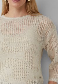 Maglione lavorato a maglia beige chiaro con una trama larga e aperta, caratterizzato da un motivo testurizzato e maniche a tre quarti. Tessuto morbido e traspirante.