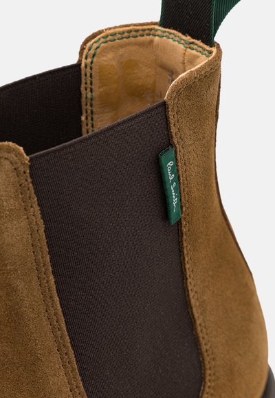 Botte Chelsea en suède marron avec un panneau élastique noir sur le côté et une étiquette de logo verte, intérieur lisse et détails de couture contrastants.