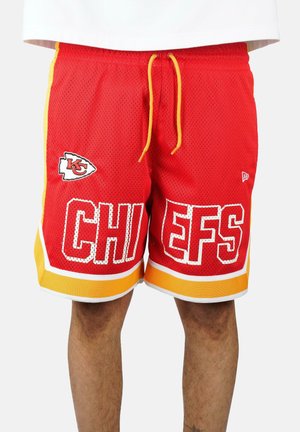 Pantaloncini sportivi rossi con rifiniture gialle con il logo e il testo dei Kansas City Chiefs, indossati da una persona in piedi davanti a uno sfondo semplice.