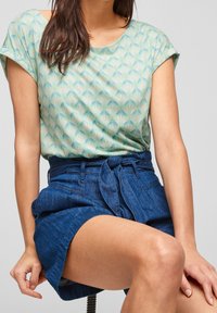 Femme portant un haut à manches courtes à motifs et une jupe en jean taille haute nouée, assise avec une main reposant sur son genou.