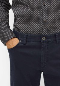 Marineblauwe broek met een gladde textuur, voorzien van een knoop- en ritssluiting, gecombineerd met een gemusterd donker shirt met knoopsluiting.