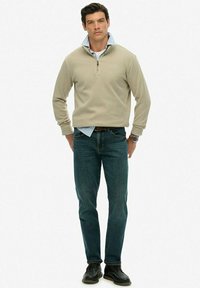 Mężczyzna w beżowym swetrze z zamkiem na ćwierć, noszący niebieską koszulę w paski, ciemne dżinsy, czarne buty i zegarek na nadgarstku, stoi z rękami za plecami.