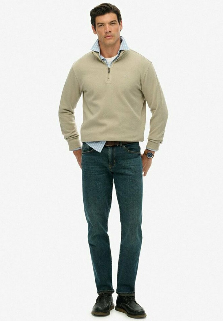 Mężczyzna w beżowym swetrze z zamkiem na ćwierć, noszący niebieską koszulę w paski, ciemne dżinsy, czarne buty i zegarek na nadgarstku, stoi z rękami za plecami.