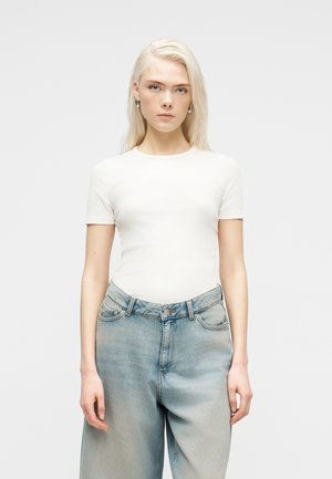 Ung kvinne med langt blondt hår iført en tettsittende hvit kortermet topp og løstsittende lyseblå jeans, stående foran en ensfarget bakgrunn.