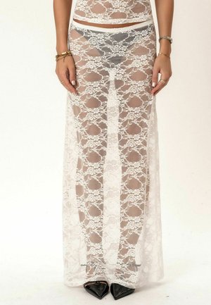 Kobieta w białej, długiej kwiecistej koronkowej spódnicy z widoczną bielizną, czarnych szpilkach z ostrym noskiem oraz złotych bransoletkach na obu nadgarstkach.