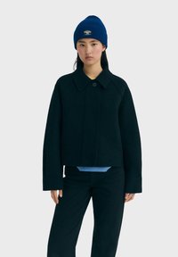 Joven mujer vestida con una chaqueta de lana oscura, pantalones a juego y un gorro de punto azul con una pequeña etiqueta en la parte frontal, de pie contra un fondo claro.