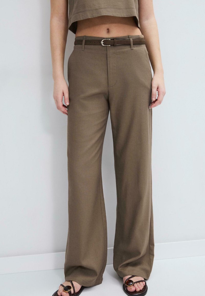 Personne portant un pantalon marron taille haute à jambes larges avec une ceinture marron assortie et des sandales noires ouvertes aux orteils, debout contre un mur blanc.