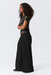 Kvinne med krøllete hår står med armene krysset, iført en mørk tie-dye T-skjorte og vide svarte bukser foran en ensfarget bakgrunn.