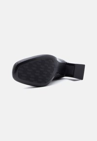 Μαύρο παπούτσι με τακούνι, εμφανιζόμενο από τη σόλα, με γεωμετρικό σχέδιο πέλματος και χοντρό τακούνι σε απλό λευκό φόντο.