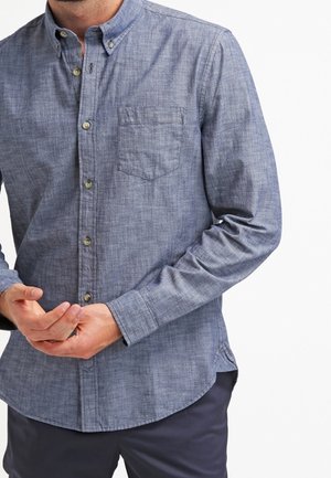 Homme portant une chemise chambray bleue à manches longues avec une poche poitrine et un pantalon foncé sur fond blanc.