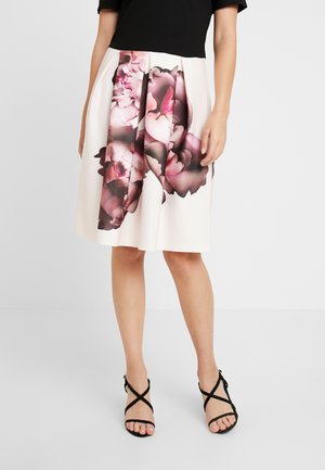 Jupe à imprimé floral crème avec des pétales roses et violets foncés ; design plissé. Portée avec un haut noir et des sandales noires à lanières.