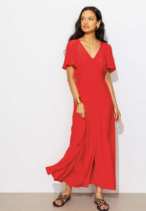 Femme aux longs cheveux foncés portant une robe rouge boutonnée, un bracelet en or et des sandales à imprimé léopard, debout contre un mur blanc.