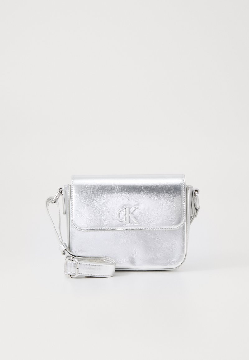 Stříbrná kovová crossbody taška s překlopným víkem, embosovaným logem, tenkým nastavitelným popruhem a hladkým povrchem. Kompaktní obdélníkový tvar.