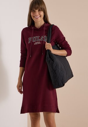 Frau lächelt, trägt ein bordeauxfarbenes Hoodie-Kleid mit dem Text "PULSE learning never stops" und hält eine große schwarze gesteppte Umhängetasche auf der Schulter.