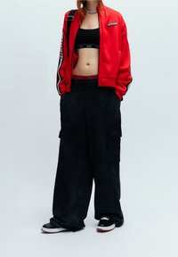 Červená zipová bunda s černými akcenty, černý crop top a volné černé cargo kalhoty. Outfit doplněn o robustní černé tenisky.