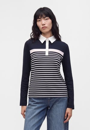 Femme aux cheveux noirs portant une chemise à rayures bleu marine et blanches à manches longues avec un col blanc et un jean bleu, debout devant un fond uni.