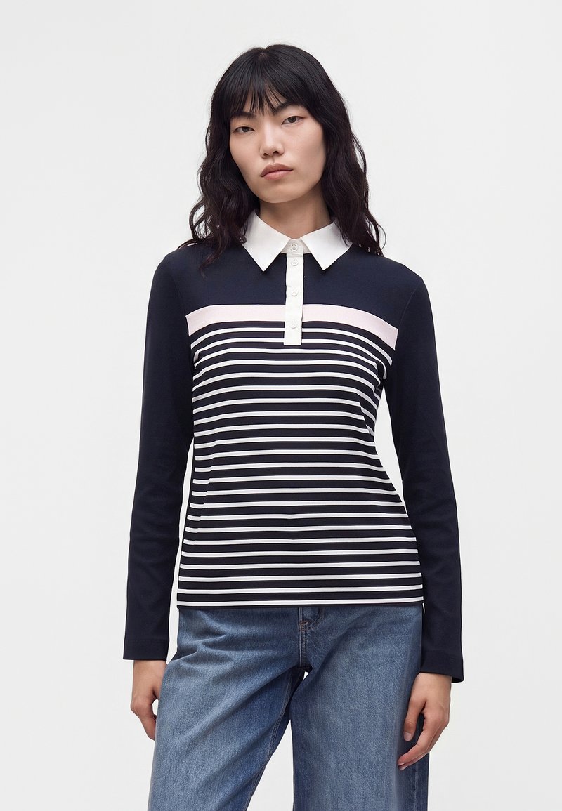 Femme aux cheveux noirs portant une chemise à rayures bleu marine et blanches à manches longues avec un col blanc et un jean bleu, debout devant un fond uni.