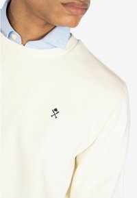 Sudadera crema con cuello redondo y un logo bordado en azul oscuro en el pecho. Debajo, se ve una camisa de color azul claro con cuello.