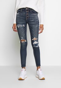 Vaqueros de mezclilla azul oscuro con corte ajustado, tiro alto, múltiples botones y detalles desgastados que incluyen desgastes en las rodillas. Combinados con zapatillas blancas.