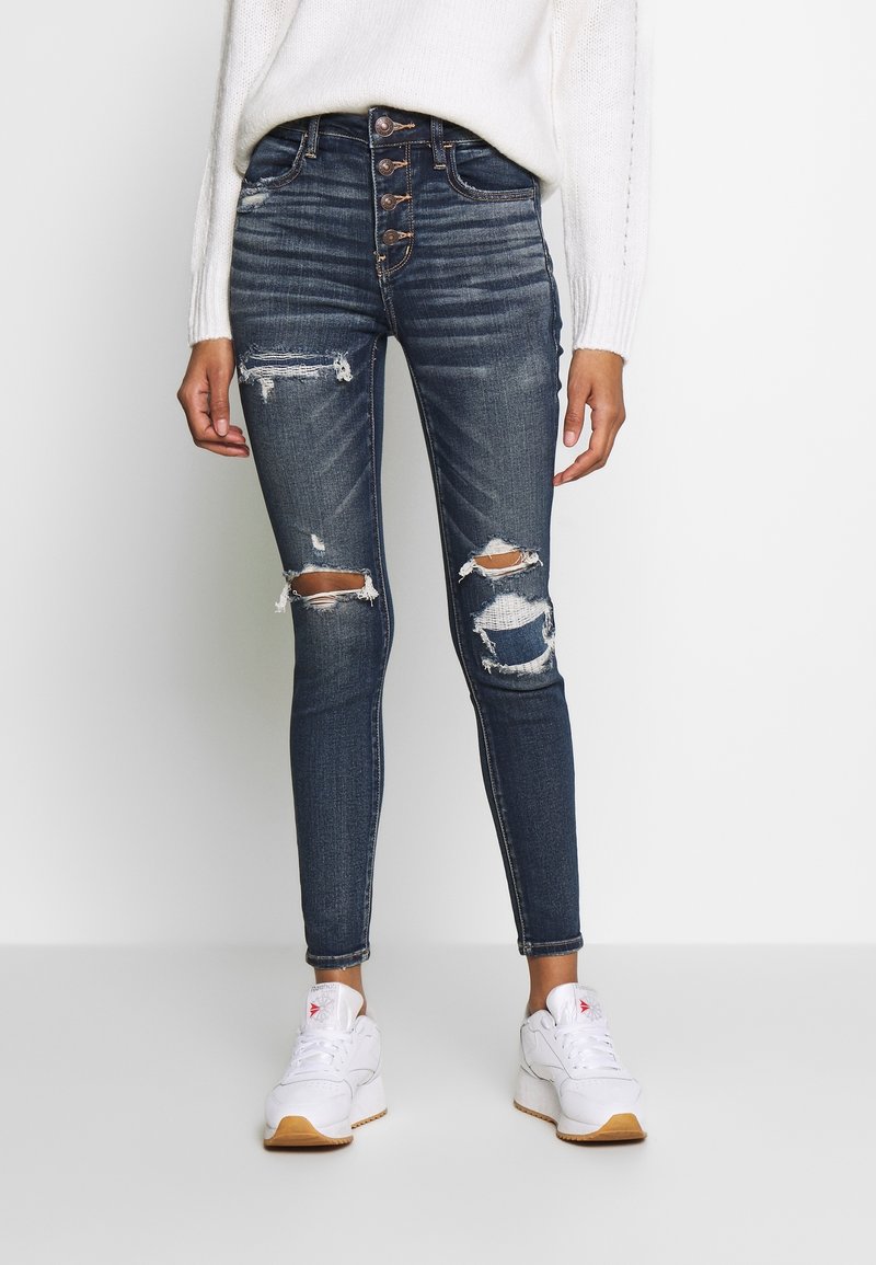 Vaqueros de mezclilla azul oscuro con corte ajustado, tiro alto, múltiples botones y detalles desgastados que incluyen desgastes en las rodillas. Combinados con zapatillas blancas.