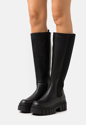 Bottes plateformes noires montantes jusqu'aux genoux avec semelles épaisses portées sur des jambes nues sur fond blanc.
