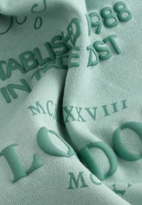 Mintu zaļa auduma ar reljefu tekstu dažādās fontu izmantojumā. Galvenās detaļas ietver "EST. 1988," "LONDON," un "MCMXXVIII" tumšāk zaļā krāsā.