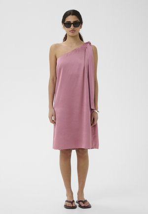 Vrouw in een knielange, éénschouder roze jurk met gedrapeerde stof, draagt zwarte zonnebril, sandalen en een wit armbandje, staand op een effen achtergrond.