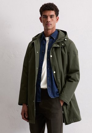 Man in een groene hooded jacket over een blauwe button-up shirt en een wit T-shirt, staand tegen een effen lichte muur.