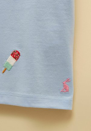 Lyseblåt stof med små broderede motiver: en flerfarvet ispind og en lyserød siddende kanin nær kanten.