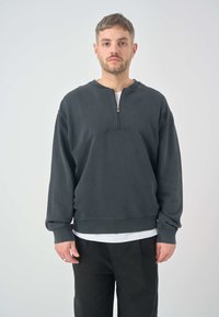 Dark gray, zippered sweatshirt with a round neckline and ribbed texture. Features wide cuffs and a relaxed fit. Paired with black trousers.