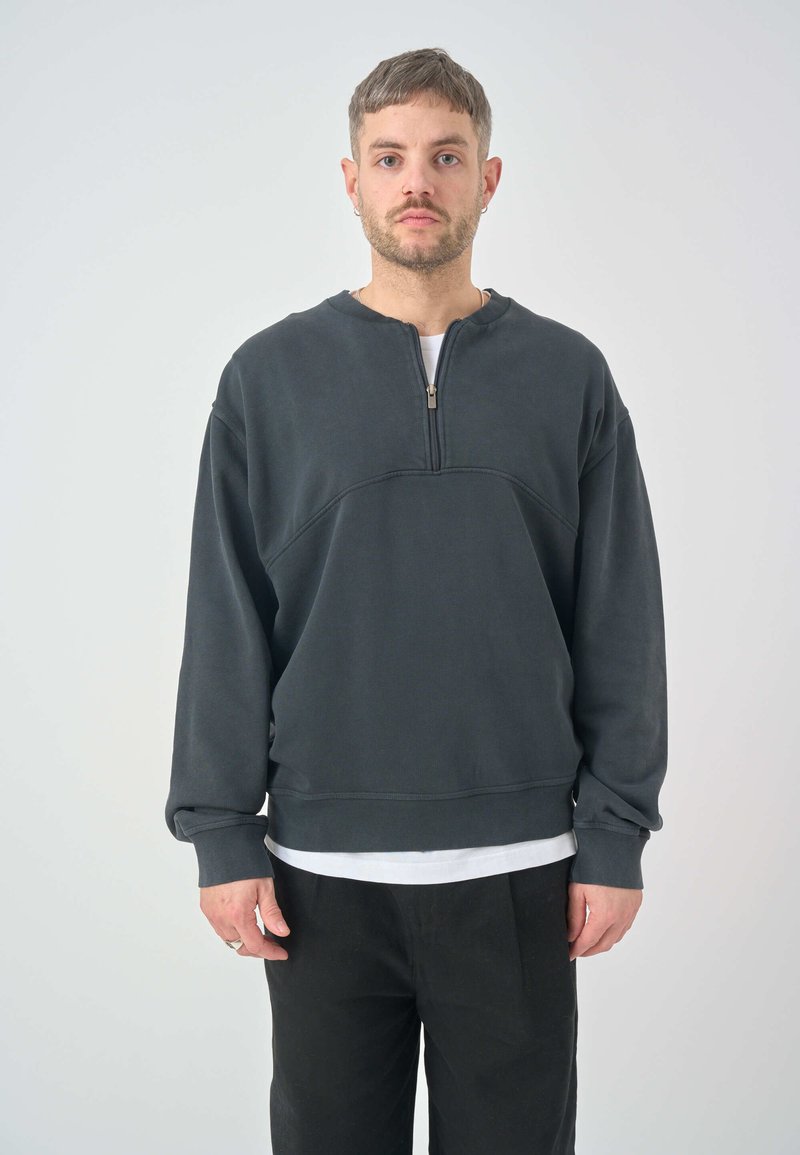 Dark gray, zippered sweatshirt with a round neckline and ribbed texture. Features wide cuffs and a relaxed fit. Paired with black trousers.