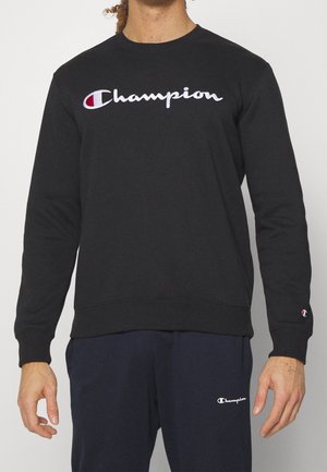 Osoba ubrana w czarną, długorękawową bluzę i granatowe spodnie, obie z białymi logo "Champion", stojąca na tle jasnego, jednolitego tła.