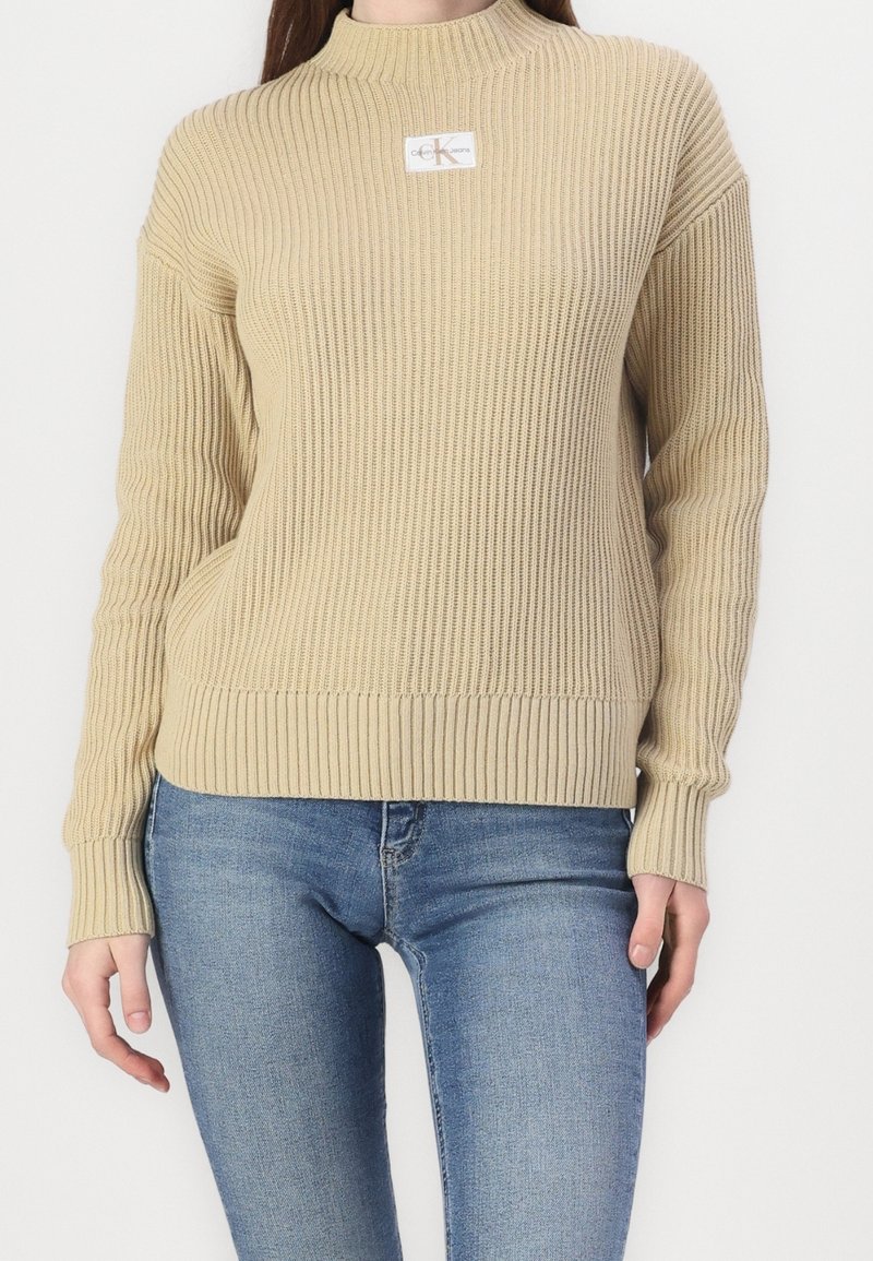 Jersey de punto beige con cuello alto, textura acanalada y parche con logo. Mangas largas con puños y dobladillo acanalados. Combinado con jeans azules.