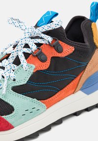 Baskets multicolores avec tissu noir surpiqué en bleu, empiècements en suède orange et menthe, semelle blanche, détail bleu au talon et lacets blancs à pois bleus.