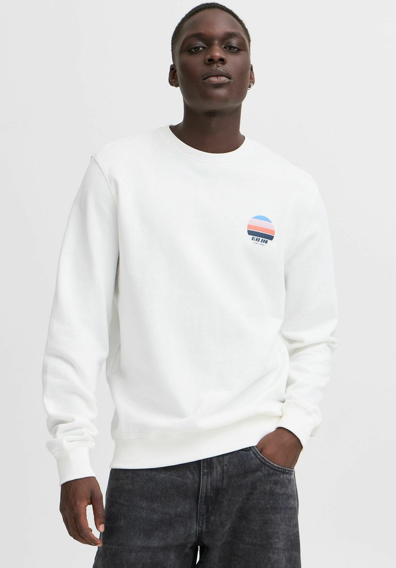 Witte sweatshirt met een ronde, kleurrijke graphic op de borst, met blauwe, roze en witte strepen. Zachte textuur, lange mouwen.