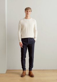 Svetar iz svetle kreme s pletenicom, temnoplave slim-fit hlače in rjave usnjene čevlje. Minimalistična zasnova, teksturirana tkanina in izpopolnjena usklajenost barv.
