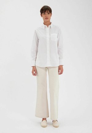 Chemise blanche à boutons avec une poche poitrine unique, manches longues et un col rigide, associée à un pantalon évasé crème et des chaussures légères à enfiler.