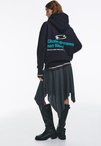 Tengerészkék kapucnis pulóver elülső kenguruzsebbel, a hátoldalán türkiz és fehér felirattal. Szürke bővülő szoknya, fekete térdig érő bőr csizma csatokkal.